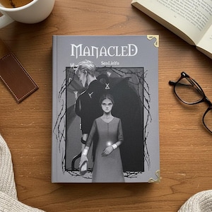 Op de afbeelding: Een boek getiteld "Manacled" met een grijswaarden illustratie van twee figuren. De cover is grijs met gouden hoekaccenten. Een kopje thee, een bladwijzer en een open boek liggen op een houten oppervlak.