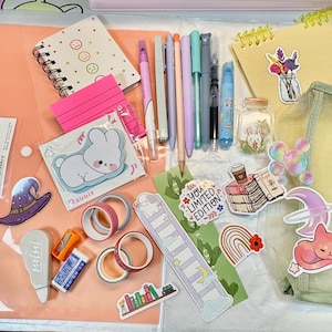 May include: A selection of stationery items including pens, stickers, washi tape, and notebooks. The items are arranged on a pink surface. Stickers feature a bunny, cat, and other cute designs. Pens are in various colours, and there is a clear pencil case.