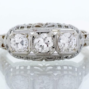 May include: A silver-toned ring featuring three round, clear, faceted stones set in a horizontal row. The band and setting are crafted with intricate, openwork metal filigree detailing that surrounds the central stones on all sides.