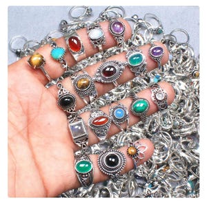 Può includere: Una collezione di anelli con pietre preziose in vari design e colori, tra cui turchese, rosso, verde e viola. Gli anelli sono incastonati in metallo argentato e mostrati su una mano, con molti altri anelli sullo sfondo.