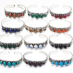 Op de afbeelding: Een verzameling zilverkleurige manchetarmbanden, elk versierd met een rij gepolijste edelstenen in verschillende kleuren. Edelstenen zijn onder meer rood, groen, blauw en oranje. De armbanden hebben een open ontwerp en decoratieve details.