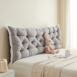 May include: A large, grey, buttoned headboard with a white border dominates the image. A stuffed bunny in a pink dress sits on the bed. A book, a mug, and a tissue box are also visible.