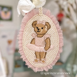 May include: An oval cross-stitch embroidery featuring a brown teddy bear wearing a pink tutu and matching hair bow. The piece is framed by a ruffled pink tulle border and a twisted rope edge, with a hanging cord and the text spiritcreaturesws.