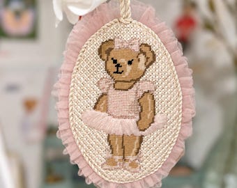 May include: An oval cross-stitch embroidery featuring a brown teddy bear wearing a pink tutu and matching hair bow. The piece is framed by a ruffled pink tulle border and a twisted rope edge, with a hanging cord and the text spiritcreaturesws.