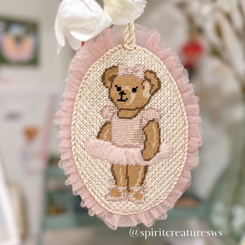 Puede incluir: Un bordado en punto de cruz ovalado que muestra un oso de peluche marr&oacute;n con un tut&uacute; rosa y un lazo a juego. La pieza est&aacute; enmarcada por un borde de tul rosa con volantes y una cuerda trenzada, con un cord&oacute;n para colgar y el texto spiritcreaturesws.