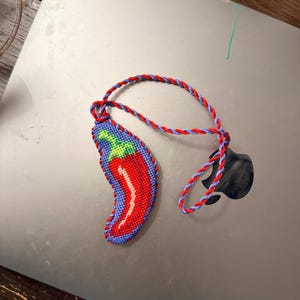 May include: A handmade chili pepper keychain. The pepper is red with a white highlight and green and blue accents. The keychain cord is red and blue. The keychain is lying on a gray surface.