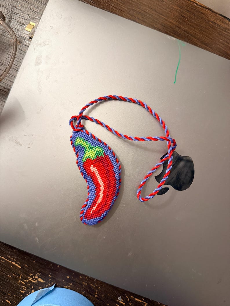 May include: A handmade chili pepper keychain. The pepper is red with a white highlight and green and blue accents. The keychain cord is red and blue. The keychain is lying on a gray surface.
