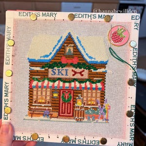 May include: A square needlepoint canvas featuring a winter ski lodge design with a snow-covered roof, a red door with a wreath, and the text SKI. The canvas is secured with gold-coloured magnets and includes a round raspberry-shaped needle minder.