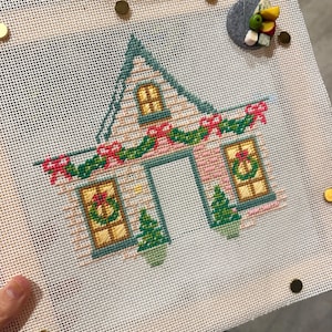 May include: Cross-stitch of a house decorated for Christmas. The house has a green roof, pink walls, and yellow windows. A garland with red bows and green leaves adorns the roofline. Two small green Christmas trees are on either side of the door.