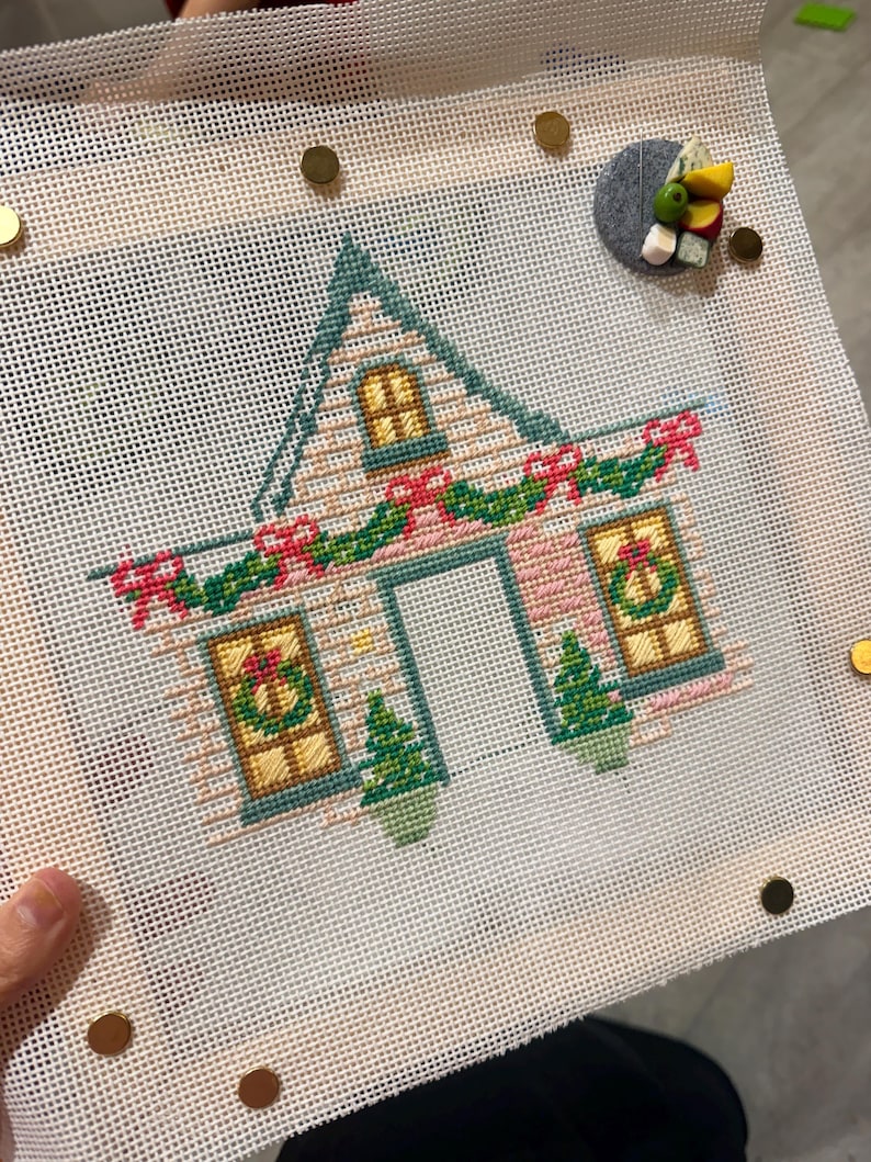 May include: Cross-stitch of a house decorated for Christmas. The house has a green roof, pink walls, and yellow windows. A garland with red bows and green leaves adorns the roofline. Two small green Christmas trees are on either side of the door.