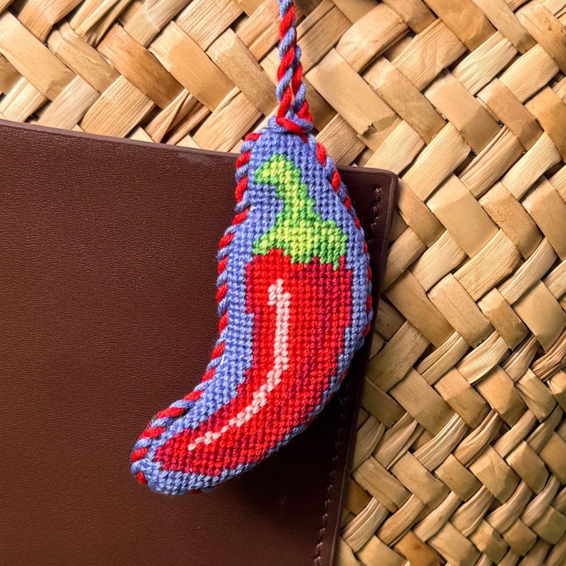 May include: A handmade, embroidered chili pepper keychain. The pepper is red with a green stem, outlined in red stitching, and set against a blue background. The keychain is attached to a red and blue twisted cord.