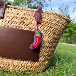 May include: A woven straw tote bag with brown leather accents and a brown leather pocket. A small, embroidered chili pepper charm with red, blue, and green details hangs from the handle. The bag sits on green grass.