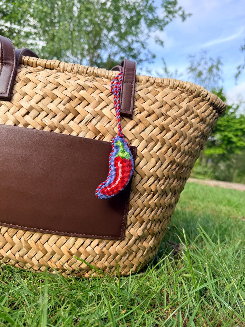 May include: A woven straw tote bag with brown leather accents and a brown leather pocket. A small, embroidered chili pepper charm with red, blue, and green details hangs from the handle. The bag sits on green grass.