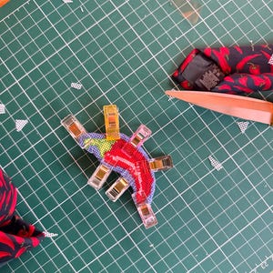 May include: A colorful, cross-stitched chili pepper design, held in place with clear clips, sits on a green cutting mat. A red and black patterned tie with chili peppers and a pair of scissors are also visible.