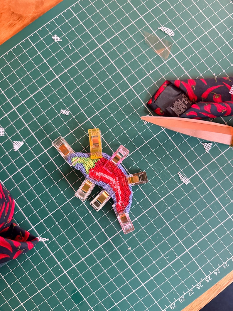 May include: A colorful, cross-stitched chili pepper design, held in place with clear clips, sits on a green cutting mat. A red and black patterned tie with chili peppers and a pair of scissors are also visible.