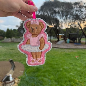 Puede incluir: Un adorno de punto de cruz con forma de osito de peluche que lleva un tut&uacute; rosa, una diadema rosa a juego y zapatillas de ballet rosas. El oso est&aacute; bordado en hilo marr&oacute;n sobre fondo rosa, con un borde de cord&oacute;n plateado y una cinta rosa.
