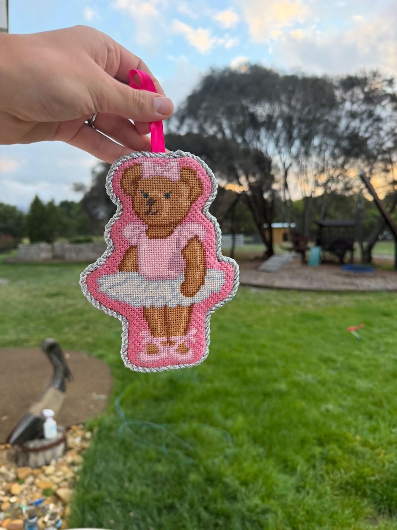 Puede incluir: Un adorno de punto de cruz con forma de osito de peluche que lleva un tut&uacute; rosa, una diadema rosa a juego y zapatillas de ballet rosas. El oso est&aacute; bordado en hilo marr&oacute;n sobre fondo rosa, con un borde de cord&oacute;n plateado y una cinta rosa.