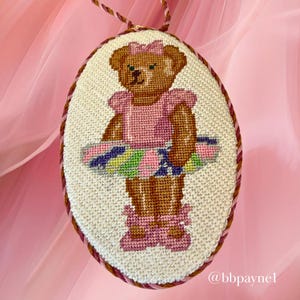 Puede incluir: Un bordado en punto de cruz ovalado que muestra un oso de peluche marr&oacute;n con un lazo rosa, una parte superior rosa y un tut&uacute; multicolor con zapatillas de ballet rosas a juego. La pieza est&aacute; enmarcada por un cord&oacute;n trenzado rosa y marr&oacute;n e incluye el texto @bbpaynel.