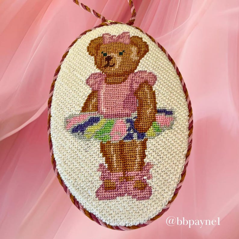 Puede incluir: Un bordado en punto de cruz ovalado que muestra un oso de peluche marr&oacute;n con un lazo rosa, una parte superior rosa y un tut&uacute; multicolor con zapatillas de ballet rosas a juego. La pieza est&aacute; enmarcada por un cord&oacute;n trenzado rosa y marr&oacute;n e incluye el texto @bbpaynel.
