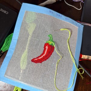 May include: Needlepoint canvas featuring a red chili pepper with a green stem, stitched on a white mesh background. The canvas is bordered with light blue fabric. Embroidery floss in green and yellow is visible, along with the numbers 86, 373, 666, 16, and 702.