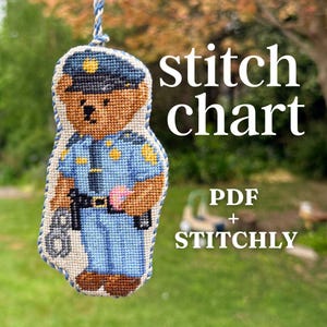 May include: A cross-stitch ornament of a brown teddy bear dressed as a police officer in a blue uniform and cap, holding handcuffs. The text stitch chart PDF + STITCHLY is visible. The ornament hangs from a braided blue and white cord.