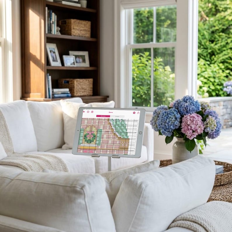 May include: A tablet displaying a cross-stitch pattern is mounted on a stand, positioned in front of a white sofa. A bouquet of blue and pink hydrangeas sits in a vase. A bookshelf and window are visible in the background.