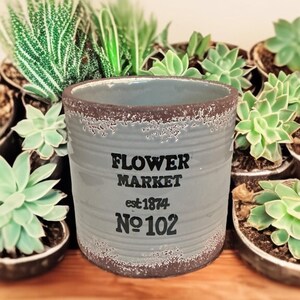 May include: A cylindrical light gray planter with a ribbed texture and a distressed brown rim. The front features black text reading FLOWER MARKET, est 1874, and No 102. The planter has a wide opening and a smooth interior surface.