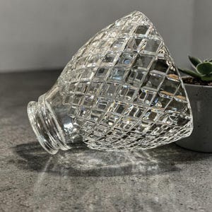 May include: A clear, diamond-cut glass lampshade with a flared base. The shade is angled, showcasing its geometric pattern and light refraction. The lampshade is a decorative lighting element.