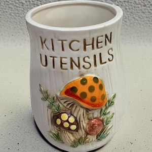 K&ouml;nnte beinhalten: Ein wei&szlig;er K&uuml;chenutensilienhalter aus Keramik in zylindrischer Form mit vertikaler Rippenstruktur. Auf der Vorderseite befindet sich der gepr&auml;gte Schriftzug KITCHEN UTENSILS &uuml;ber einem farbigen Relief aus drei Pilzen und gr&uuml;nem Blattwerk.