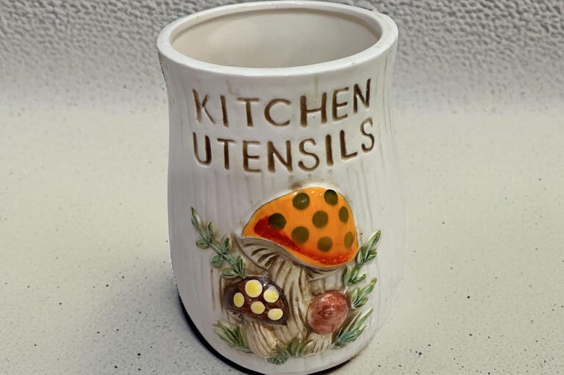 K&ouml;nnte beinhalten: Ein wei&szlig;er K&uuml;chenutensilienhalter aus Keramik in zylindrischer Form mit vertikaler Rippenstruktur. Auf der Vorderseite befindet sich der gepr&auml;gte Schriftzug KITCHEN UTENSILS &uuml;ber einem farbigen Relief aus drei Pilzen und gr&uuml;nem Blattwerk.
