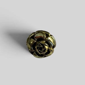 May include: A metallic gold-toned button shaped like a blooming rose. The object features layered, curved petals that spiral toward a central point, creating a dimensional, floral appearance with a polished, reflective surface finish.