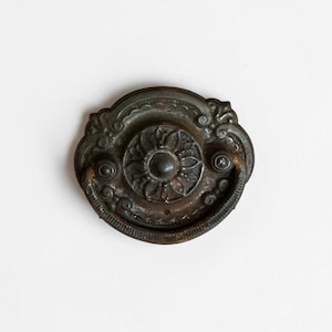 May include: A dark bronze-toned metal drawer pull featuring a circular backplate with an embossed floral pattern and a curved, swinging handle. The hardware displays a weathered, aged patina with intricate scrollwork details along the outer edge.