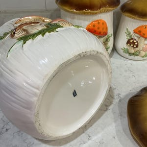 K&ouml;nnte beinhalten: Ein Set aus drei pilzf&ouml;rmigen Keramikdosen mit wei&szlig;en K&ouml;rpern und braunen Deckeln. Die Dosen zeigen erhabene, handbemalte orangefarbene und braune Pilzdetails mit gr&uuml;nem Blattwerk. Auf dem Boden einer Dose befindet sich ein kleiner blauer Aufkleber mit der Aufschrift JAPAN.