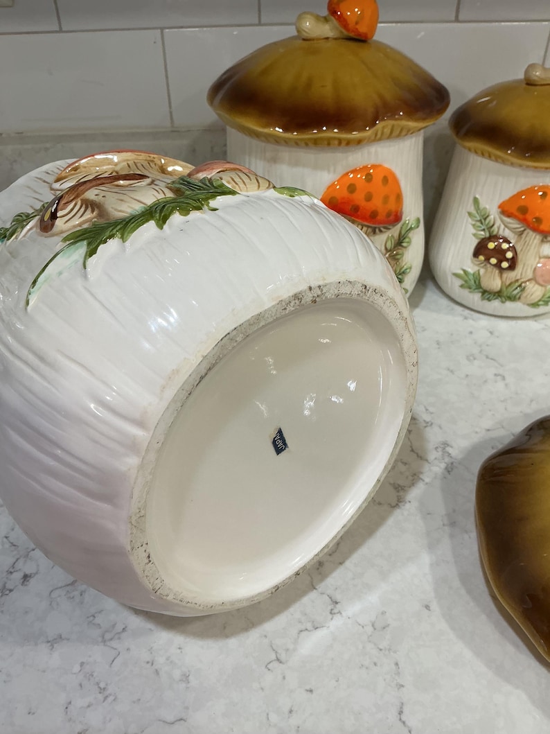 K&ouml;nnte beinhalten: Ein Set aus drei pilzf&ouml;rmigen Keramikdosen mit wei&szlig;en K&ouml;rpern und braunen Deckeln. Die Dosen zeigen erhabene, handbemalte orangefarbene und braune Pilzdetails mit gr&uuml;nem Blattwerk. Auf dem Boden einer Dose befindet sich ein kleiner blauer Aufkleber mit der Aufschrift JAPAN.