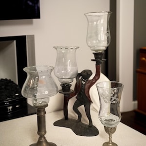 May include: An assortment of decorative candle holders. The centerpiece is a whimsical sculpture of a rabbit in a suit holding a candle holder. Other holders are made of metal and glass, with clear glass shades. The holders vary in height and design.