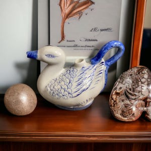 May include: A white ceramic duck teapot with blue and white painted details. The teapot has a blue handle and a spout shaped like a duck's beak. The teapot is sitting on a wooden shelf.
