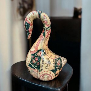 May include: A decorative swan figurine featuring a decoupage-style finish with a patchwork pattern of cream, dark green, and red floral motifs. The swan has a curved neck and a smooth, glossy surface, showcasing intricate botanical illustrations.