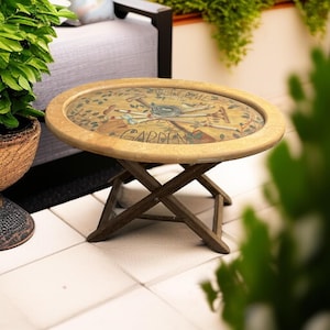 May include: A round wooden side table with a light brown frame and a central glass panel. The surface features a painted illustration of garden tools, a bird nest, and the text "WELCOME TO THE GARDEN" in black script. It has a folding X-shaped base.