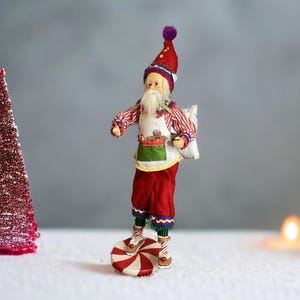 May include: A whimsical Christmas elf figurine, dressed in a red and white striped shirt, red pants, and a matching hat with a purple pom-pom. The elf stands on a red and white peppermint swirl base, carrying a sack and apron with gingerbread cookies.