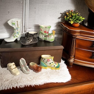 May include: A collection of miniature ceramic shoes and boots in various styles and colors. The collection includes a white boot with blue floral accents, a black boot, and a green boot with red flowers. Other shoes include a beige sandal, a white shoe with pink flowers, and a brown clog.