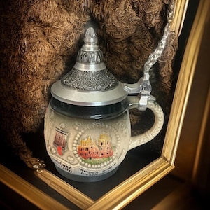 May include: A ceramic beer stein featuring a metallic lid with an ornate embossed pattern and a hinged handle. The body is cream-colored with a relief illustration of a building and the German text "WINZIG AM DURSTE RIESENGROSS".