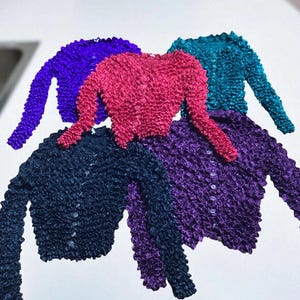 May include: A collection of five textured, long-sleeved cardigans in various colors: purple, magenta, teal, navy, and dark purple. Each cardigan has a button-up front and a unique, ruffled texture.