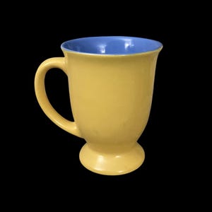 May include: A yellow ceramic mug with a blue interior. The mug has a wide base and a handle.