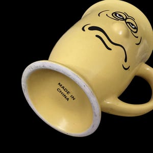 May include: Yellow ceramic mug with a cartoon face and a frown. The mug has a handle and says "Made in China" on the bottom.