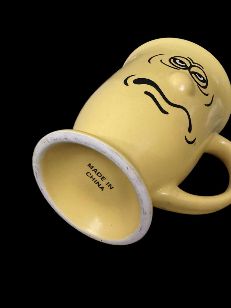 May include: Yellow ceramic mug with a cartoon face and a frown. The mug has a handle and says "Made in China" on the bottom.