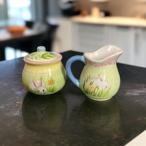 May include: A ceramic sugar bowl with a lid and a creamer pitcher. Both are painted with a light green and yellow background, featuring a white bunny, flowers, and grass. The creamer has a blue handle and pink trim.