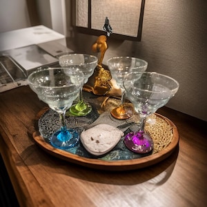 May include: Four clear margarita glasses with colored stems (blue, green, orange, and purple) sit on a wooden tray with a decorative stone. A gold sculpture with a butterfly is in the background. The tray is on a wooden surface.
