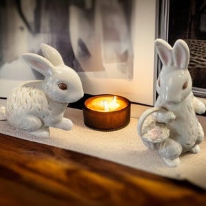 May include: Two white ceramic bunny figurines with pink-lined ears and brown eyes sit on a shelf. A lit candle in a wooden holder is between them. The bunnies are part of a spring or Easter home decor theme.