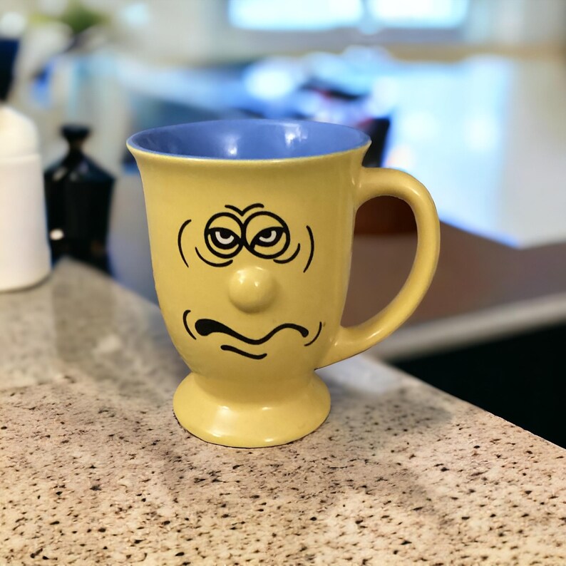 May include: Yellow ceramic coffee mug with a blue rim and a cartoon face with a grumpy expression. The mug has a small, round nose and a black, frowning mouth.