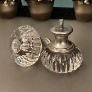 May include: Two clear glass curtain finials with a silver metal base. The finials have a ribbed design and are shaped like a rounded dome.